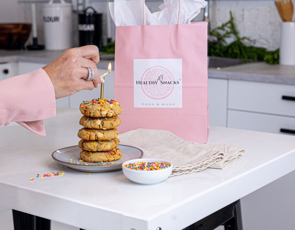 Stack of cookies with a lit candle on a plate, next to a pink bag labeled 'Healthy Snacks' in a kitchen setting.
